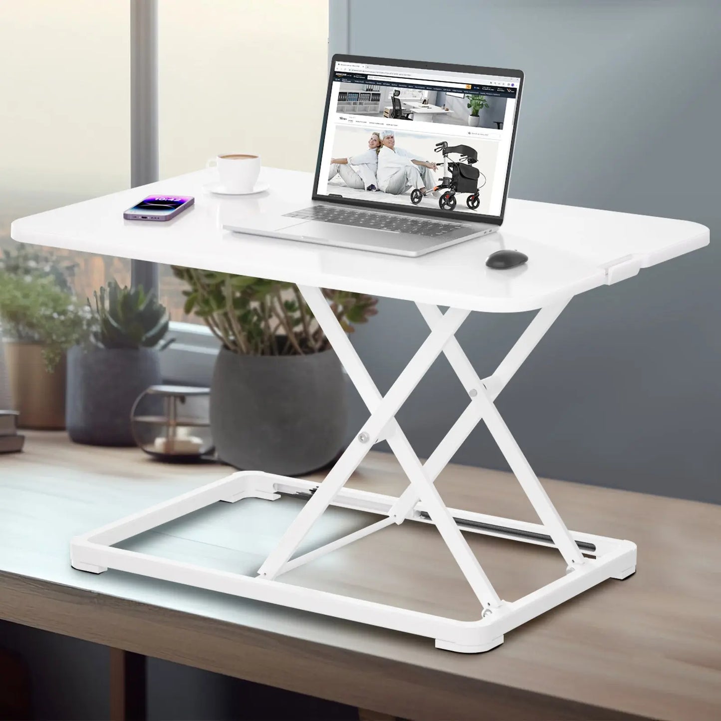 A white standing desk displayed in a home office setting near a window, complete with a laptop, smartphone, and houseplants decorating the space.