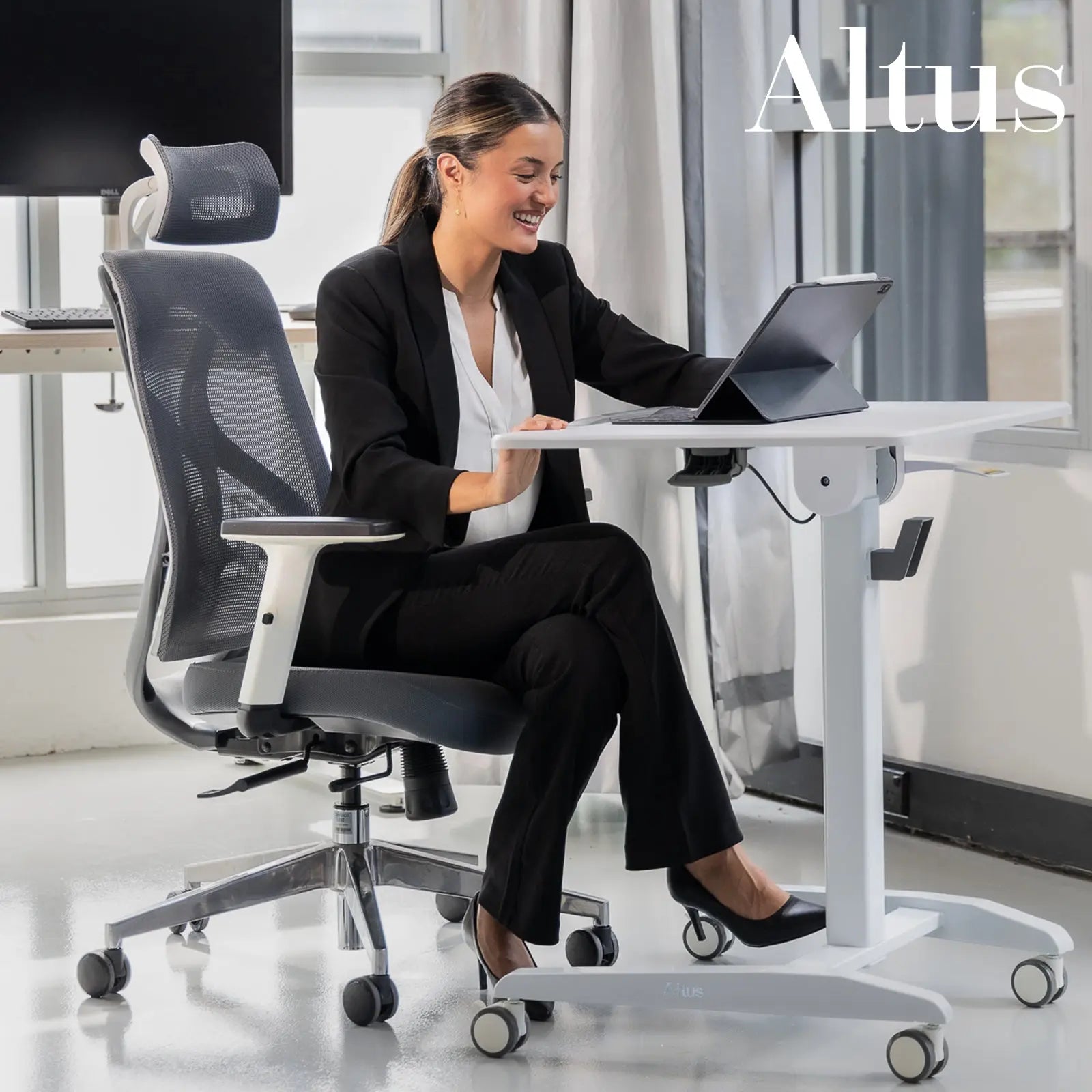 Smiling woman in a business suit adjusting a white sit-stand desk in a modern office setting.