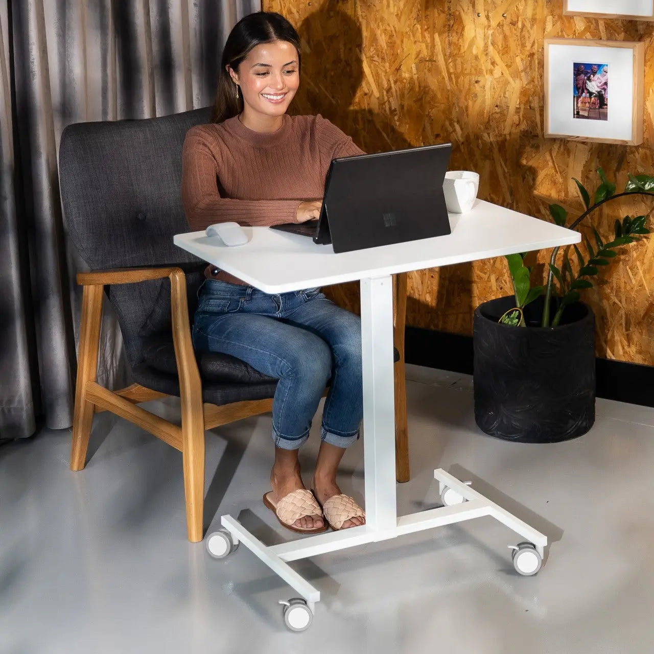 Young woman smiling while using a laptop on a white mobile desk in an office setting with a rustic wood panel backdrop and a lush potted plant.