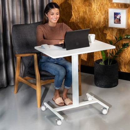 Young woman smiling while using a laptop on a white mobile desk in an office setting with a rustic wood panel backdrop and a lush potted plant.