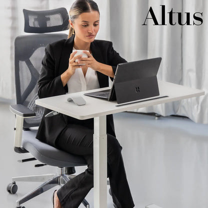 A professional setting with a woman seated at a modern, white standing desk, working on a laptop, with a neutral-toned office chair and background.