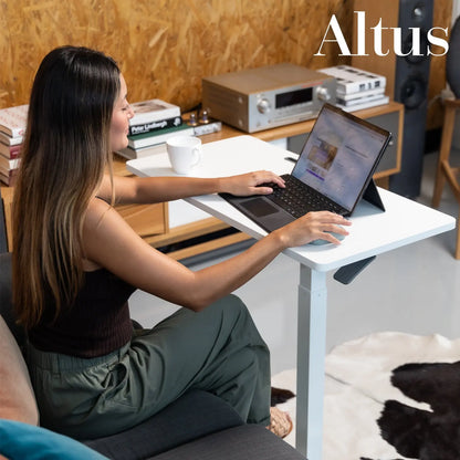 Image of a young woman seated at a white standing desk in a casual setting, using a laptop.