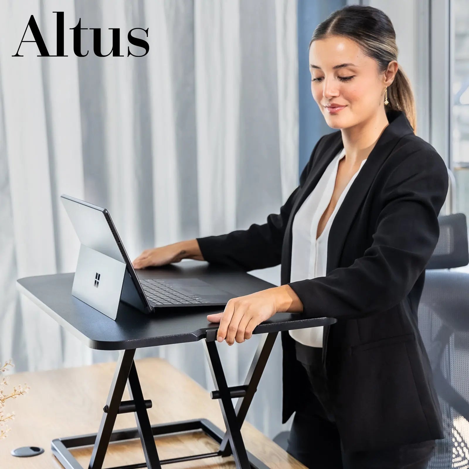 Close-up view of a woman using a laptop on a black sit-stand desk converter in a modern office setting.