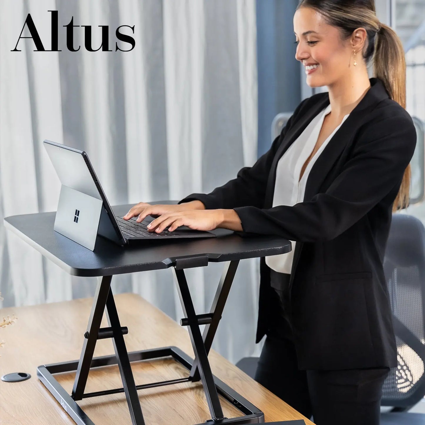 A professional woman in a business suit working at a standing desk riser in an office environment. She is typing on a laptop with a focused expression.