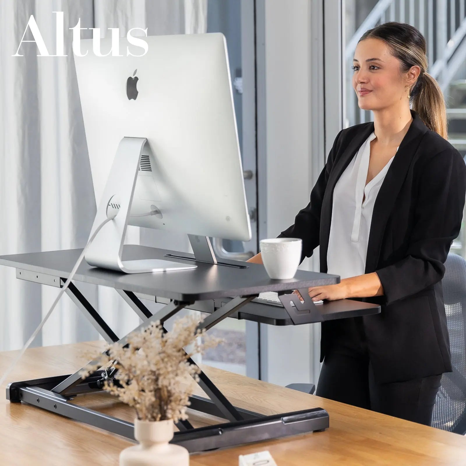 Woman using a black desk riser in a sleek office setting, adjusting the height with a lever, next to a modern glass window.