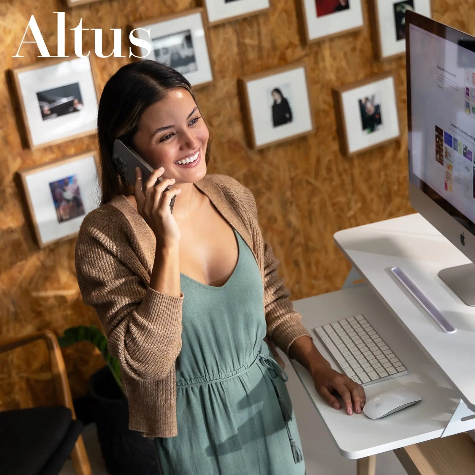 A woman in a casual outfit smiling while talking on the phone and working at an adjustable white desk converter in a cozy office environment.