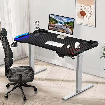 A setup showing the same height-adjustable desk in a home office environment with a computer, keyboard, and various desk accessories. The background includes a white wall and a framed abstract art piece.